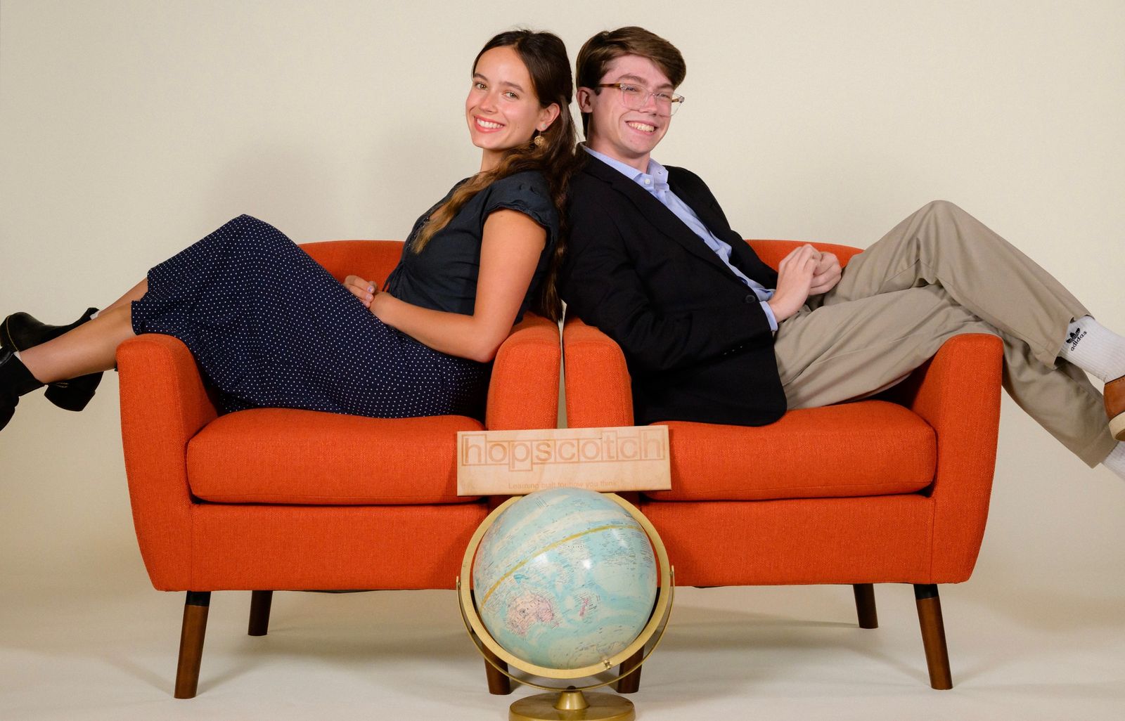 Lily Bryan and Liam Hyde, Hopscotch co-founders, sitting back to back on an orange sofa beside a globe